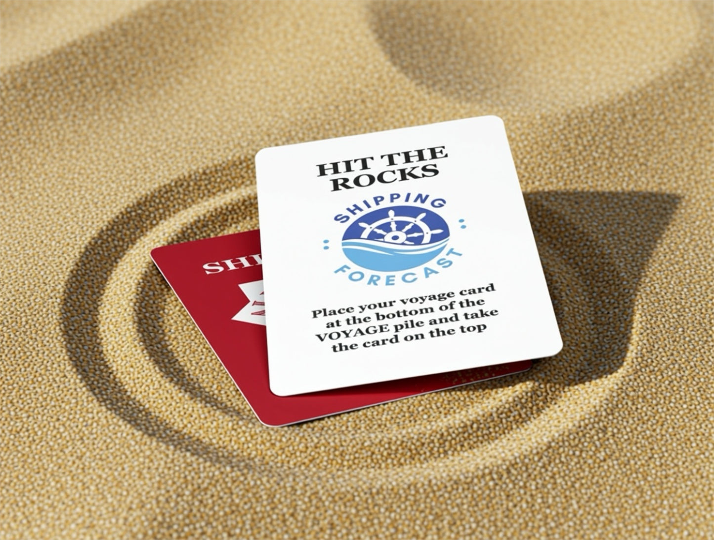Two playing cards on sand, one with 'Hit the Rocks' text and a ship icon, the other red.