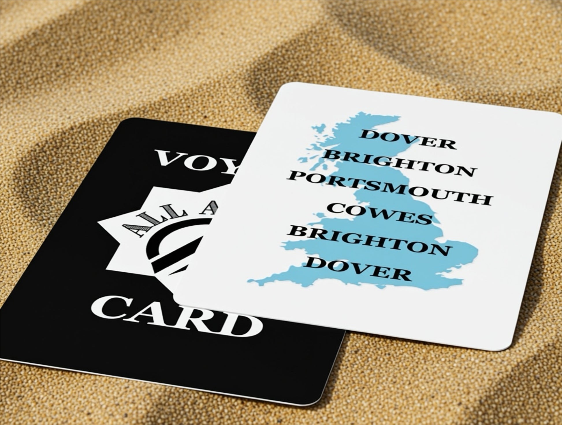 Two playing cards on a sandy surface, one black with text and logo, the other white with blue map and text.