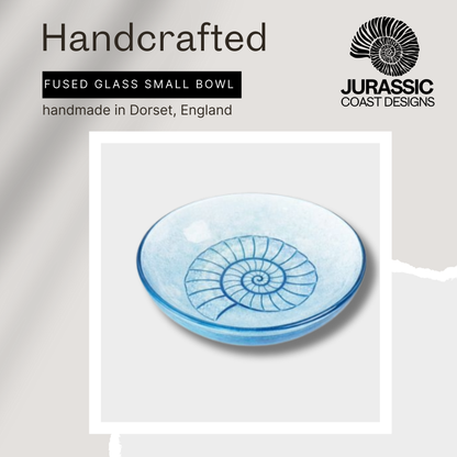 Handcrafted fused glass small bowl with ammonite design, branded 'Jurassic Coast Designs', on a light gray background.