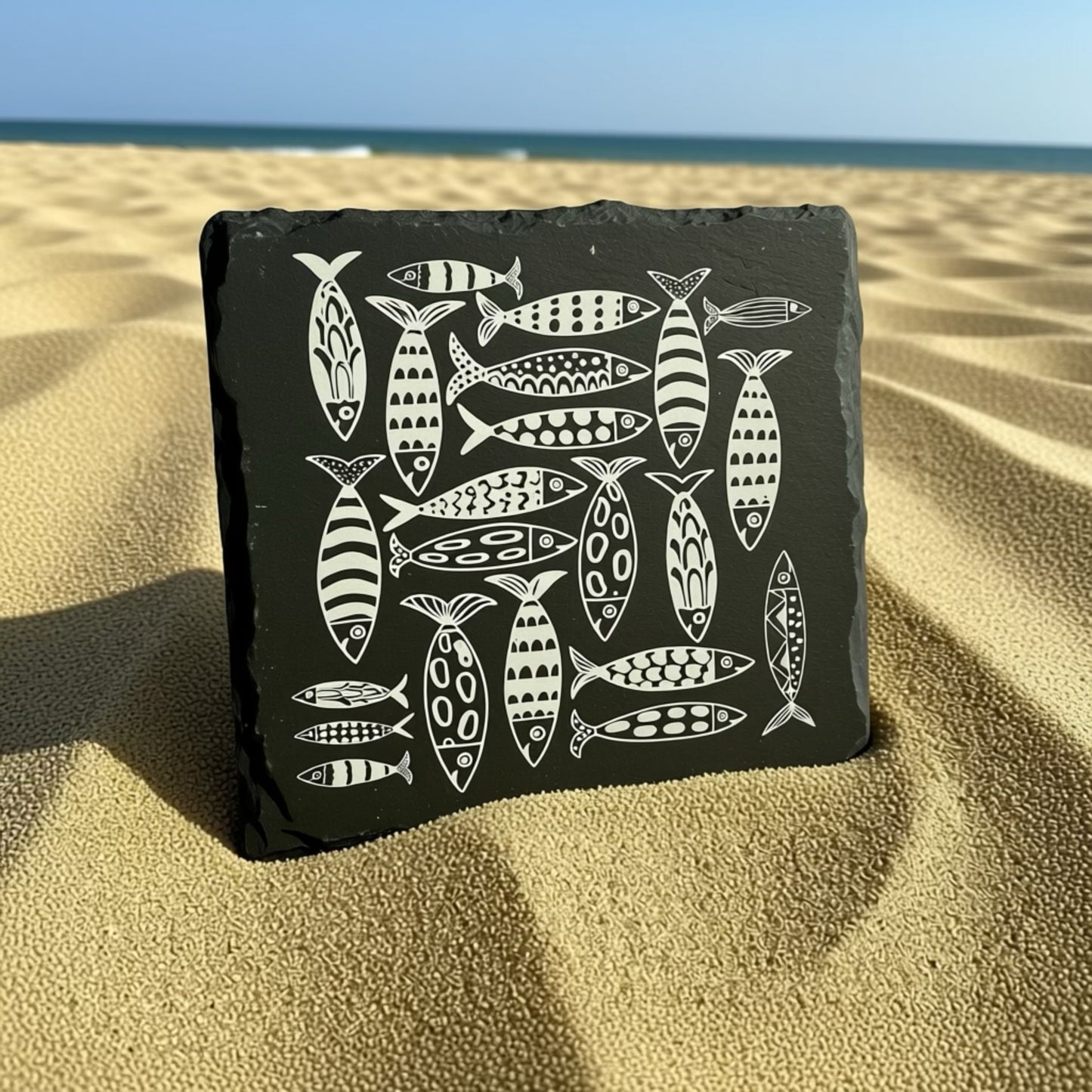 Black slate coaster with white fish illustrations on a sandy beach