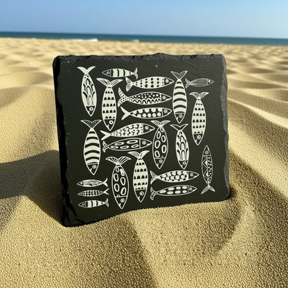 Black slate coaster with white fish illustrations on a sandy beach