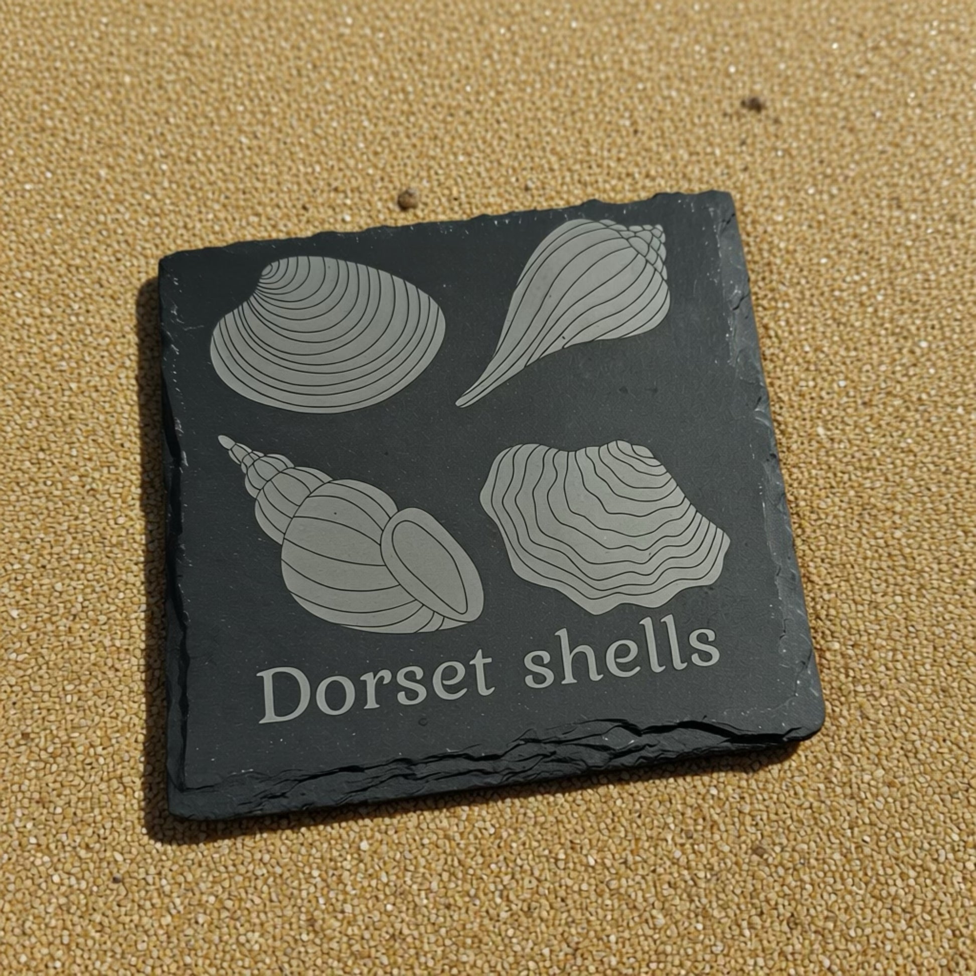 Slate coaster with engraved shell designs and 'Dorset shells' text on a sandy background
