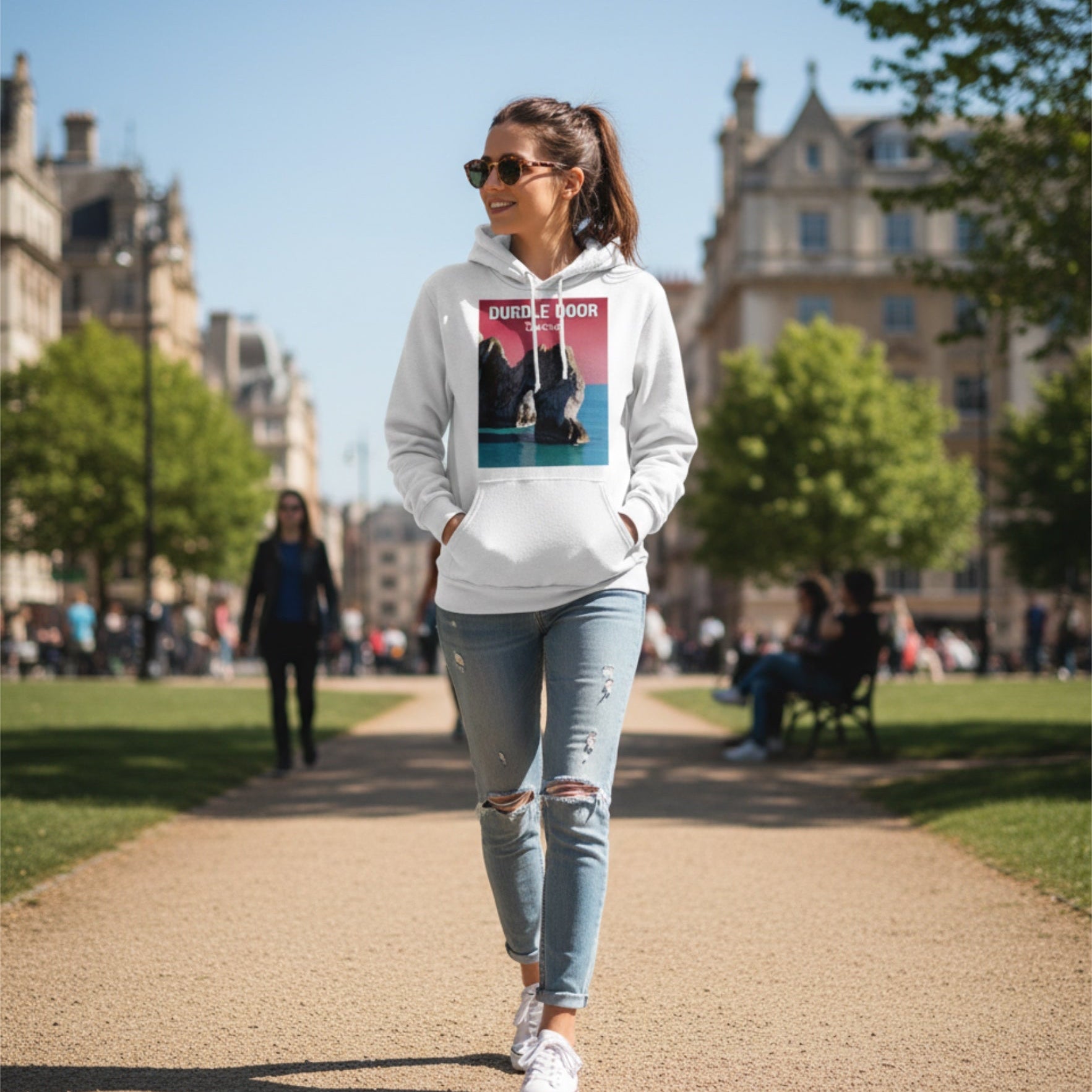 Person wearing a hoodie with a graphic Durdle Door design, walking in an outdoor setting with buildings and trees in the background.