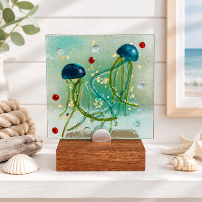 A square glass art piece featuring two blue jellyfish with green tentacles inside a translucent aqua glass panel, mounted on a rectangular wooden base.