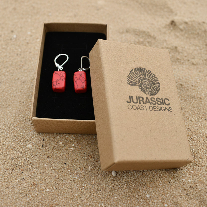 Red earrings in an open box with 'Jurassic Coast Designs' branding on a light background
