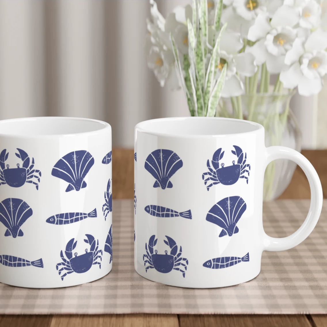 Two white mugs with blue sea-themed designs on a wooden table with a checkered tablecloth.