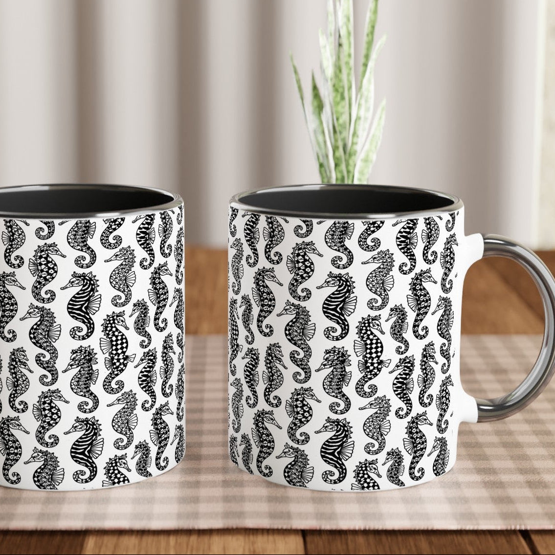 Two mugs with seahorse pattern on a wooden table with a plant in the background.